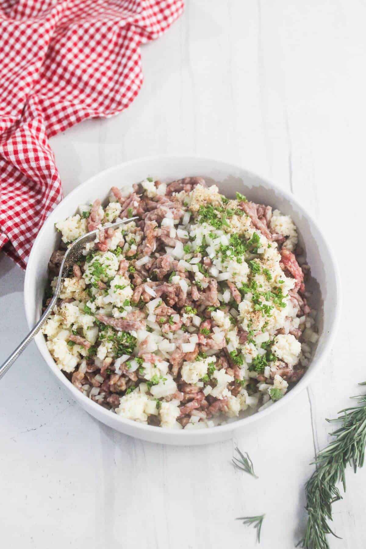 A white bowl filled with a mixture of ground meat, chopped onions, and herbs, with a spoon inside, placed on a white surface next to a red checkered cloth.