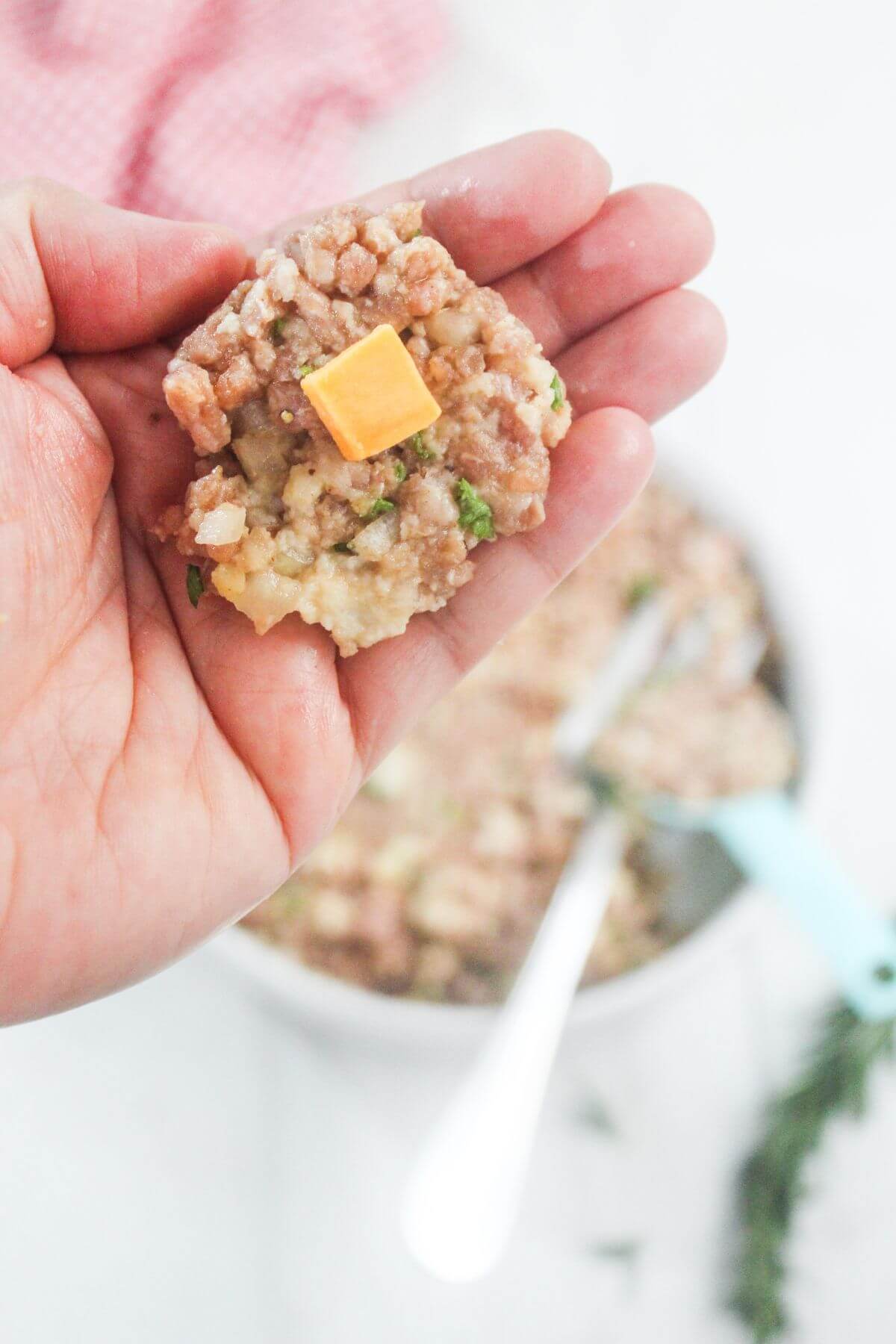 A hand holds a portion of uncooked stuffing mixture topped with a small cube of cheddar cheese, with a mixing bowl and utensils in the background.