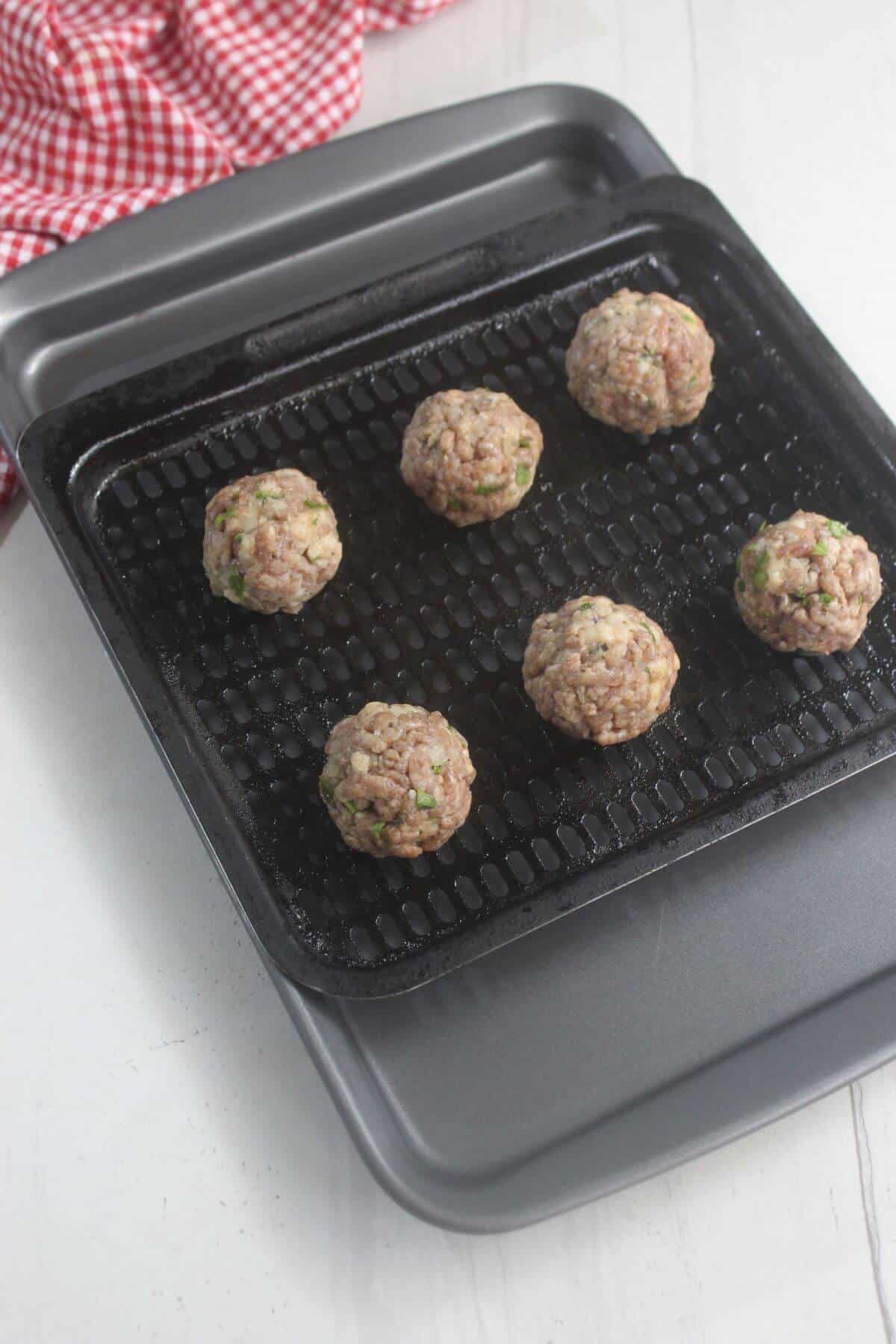 Six raw meatballs arranged on a black air fryer tray placed on a baking sheet, with a red checkered cloth in the background.