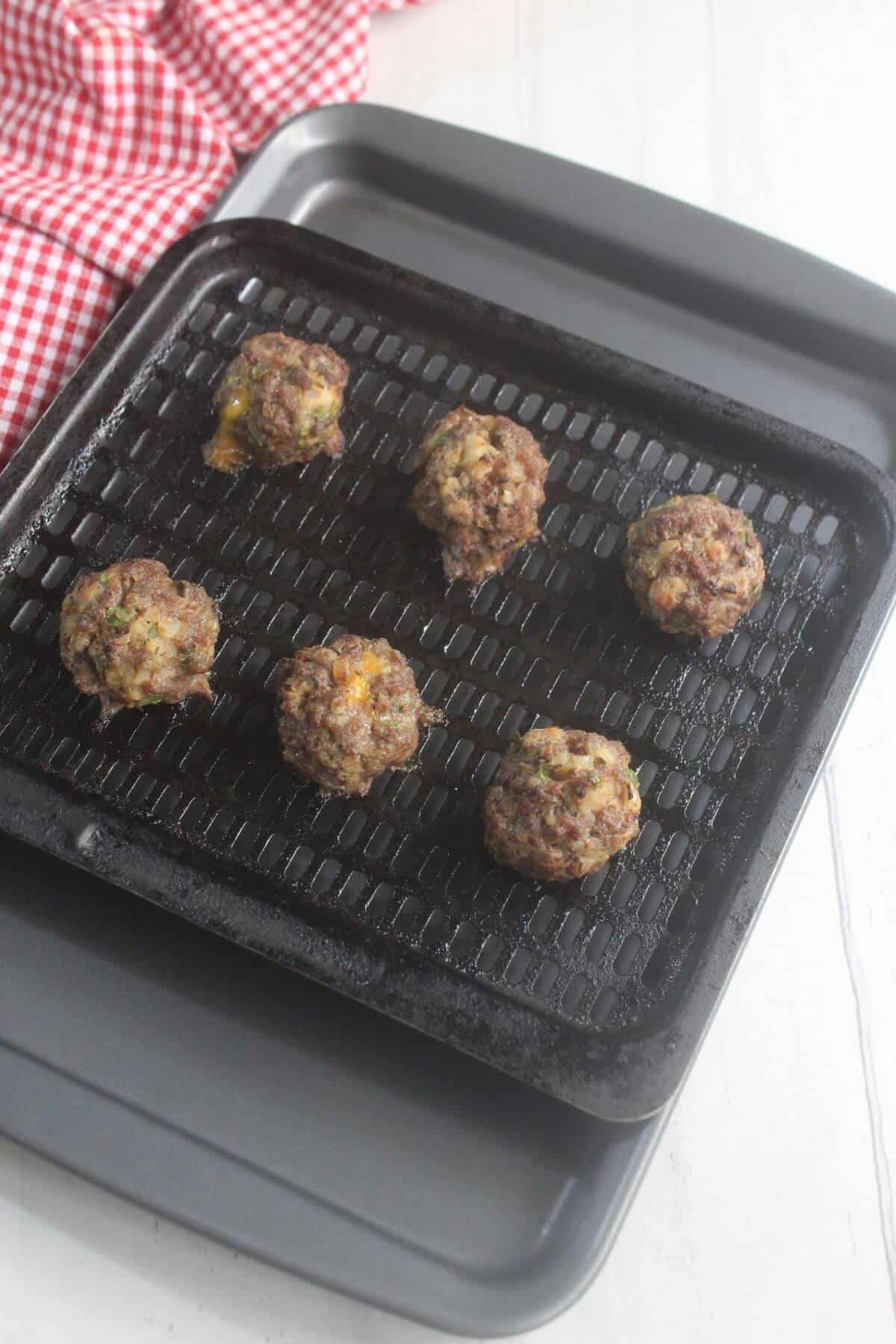 Five cooked meatballs on a black grill rack over a baking tray, with a red and white checkered cloth nearby on a white surface.