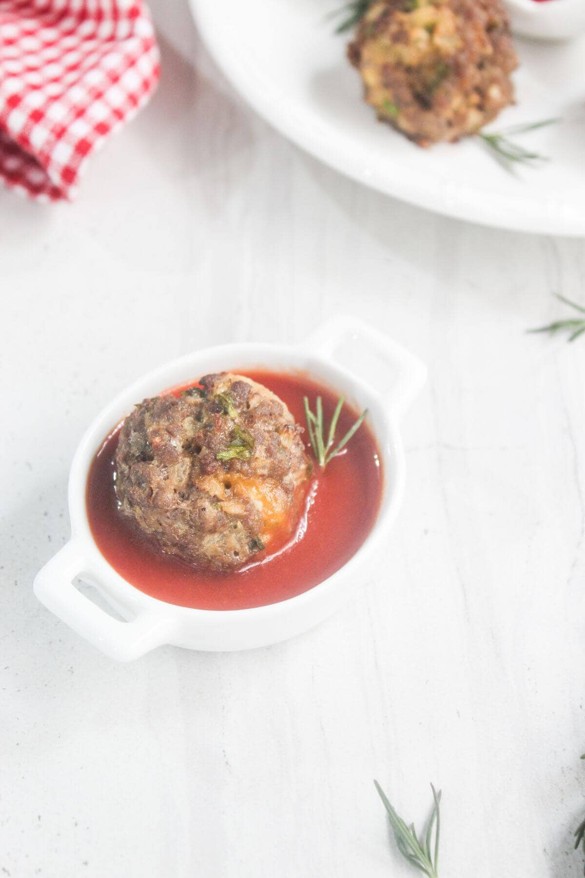 A single meatball in tomato sauce garnished with rosemary, served in a small white dish on a light surface.