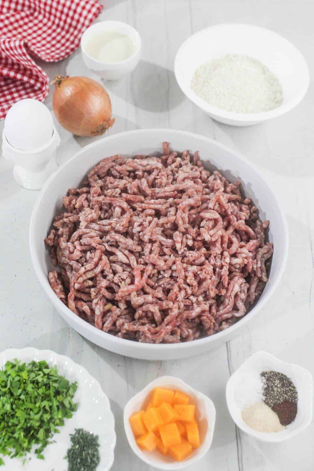Ground meat in a bowl surrounded by small bowls of chopped herbs, cheese cubes, seasonings, rice, milk, an onion, and an egg on a white surface.