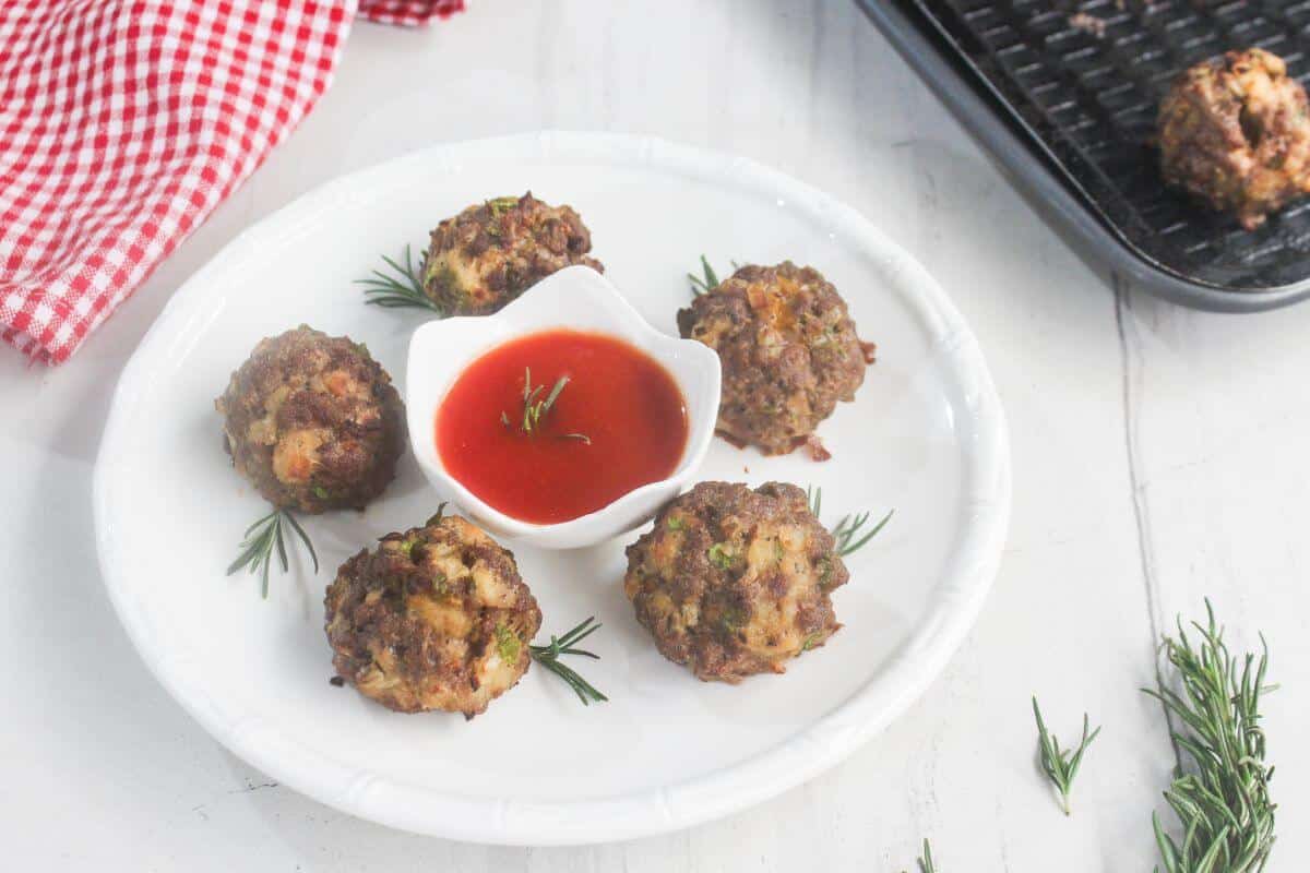 Five meatballs are arranged on a white plate with a small bowl of red dipping sauce in the center, garnished with rosemary sprigs. A red checkered cloth is nearby.