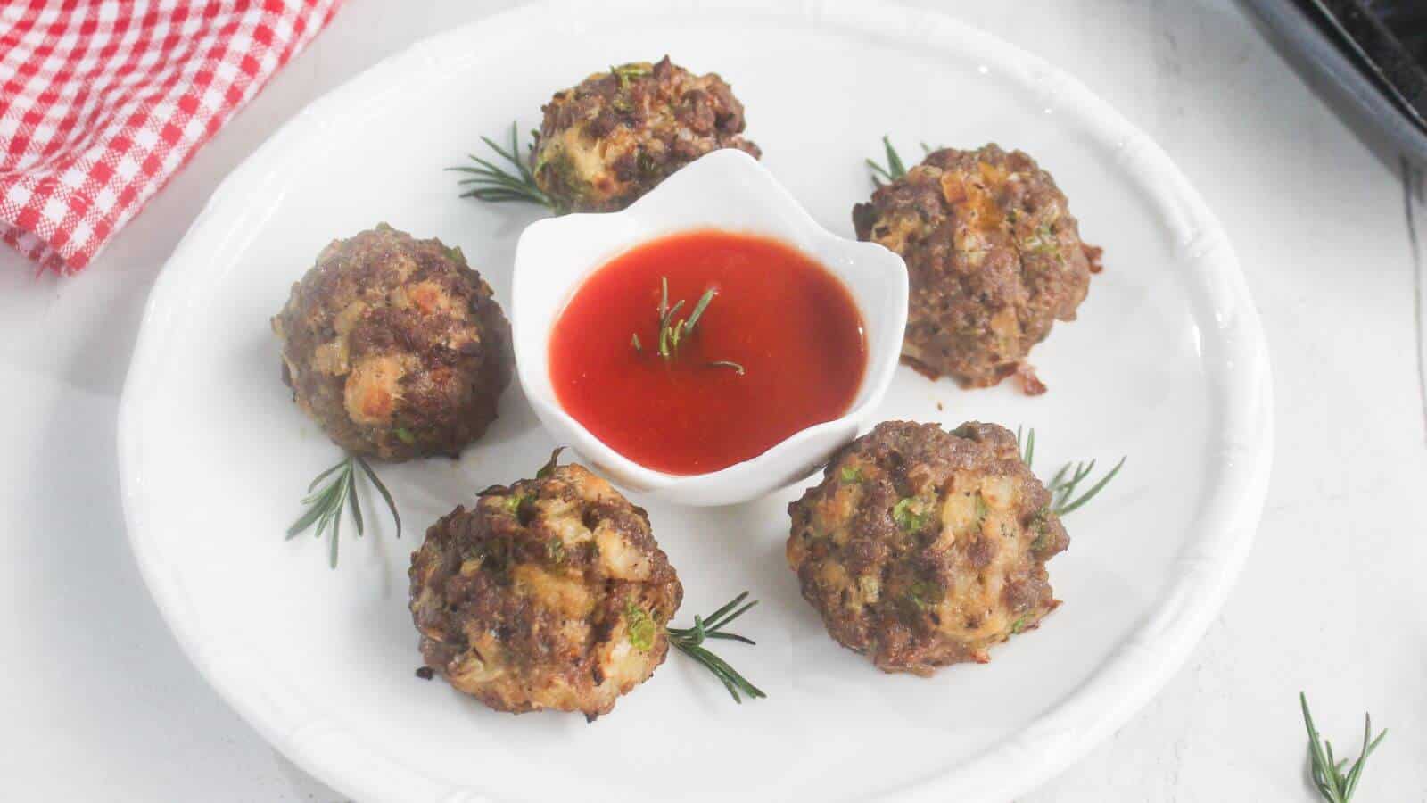 Five meatballs arranged around a small bowl of red dipping sauce on a white plate, garnished with sprigs of rosemary.