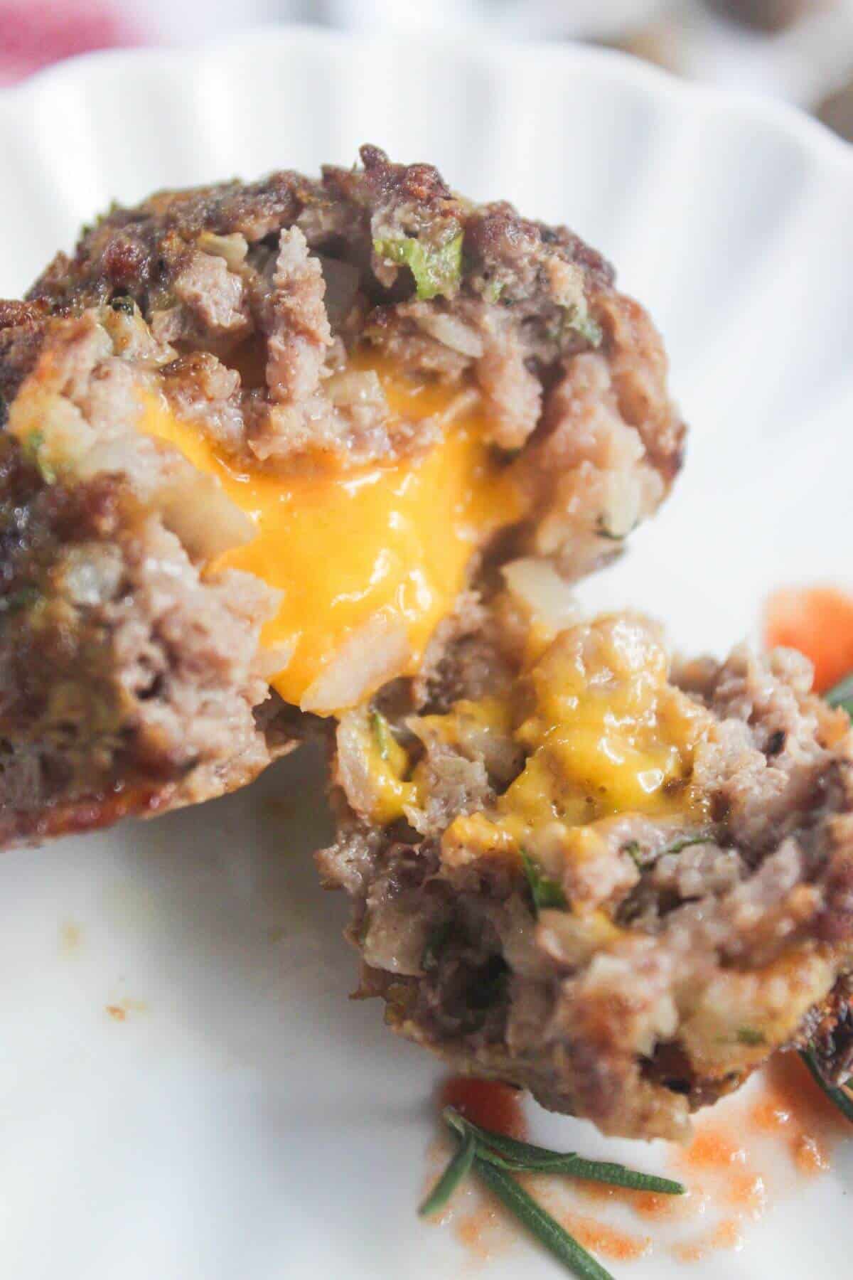 A stuffed meatball cut open to reveal melted cheddar cheese inside, served on a white plate with a sprig of rosemary and a small amount of sauce.