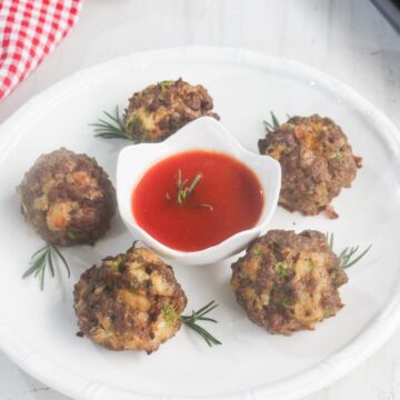 Five meatballs are arranged on a white plate with rosemary sprigs, surrounding a small dish of red dipping sauce in the center.