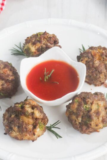 Five meatballs are arranged on a white plate with rosemary sprigs, surrounding a small dish of red dipping sauce in the center.