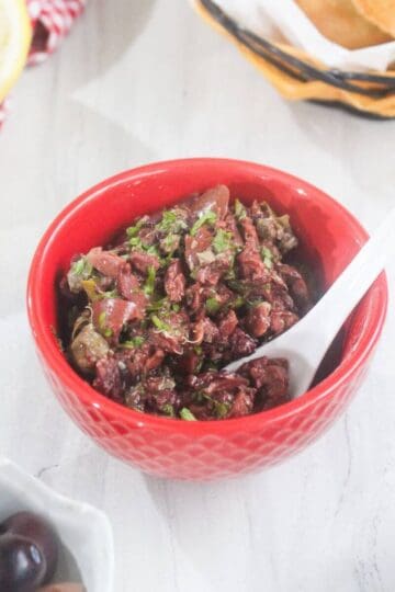 A red bowl filled with chopped olive tapenade and a white spoon, surrounded by lemons, olives, and a basket of bread on a white surface.