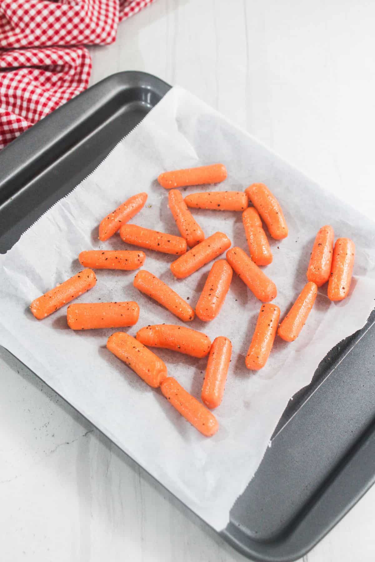 Baby carrots seasoned with pepper are spread on a parchment-lined baking tray, ready for roasting. A red and white checkered cloth is in the top left corner.