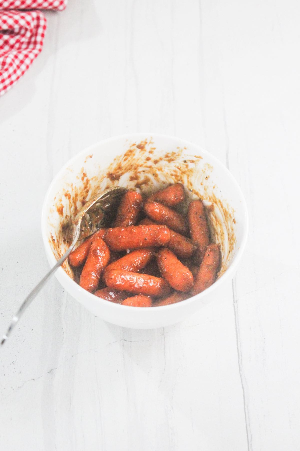 A white bowl filled with cocktail sausages coated in sauce, with a spoon inside, placed on a light-colored countertop.