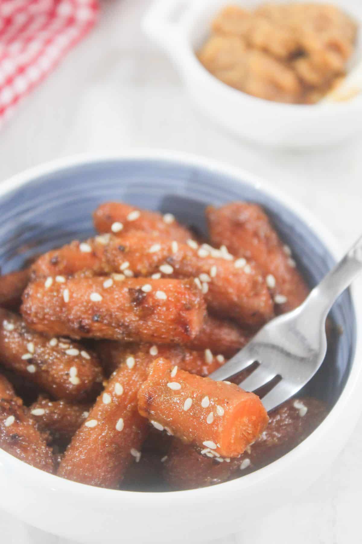 A bowl of glazed carrots topped with sesame seeds, with a fork and another dish in the background.