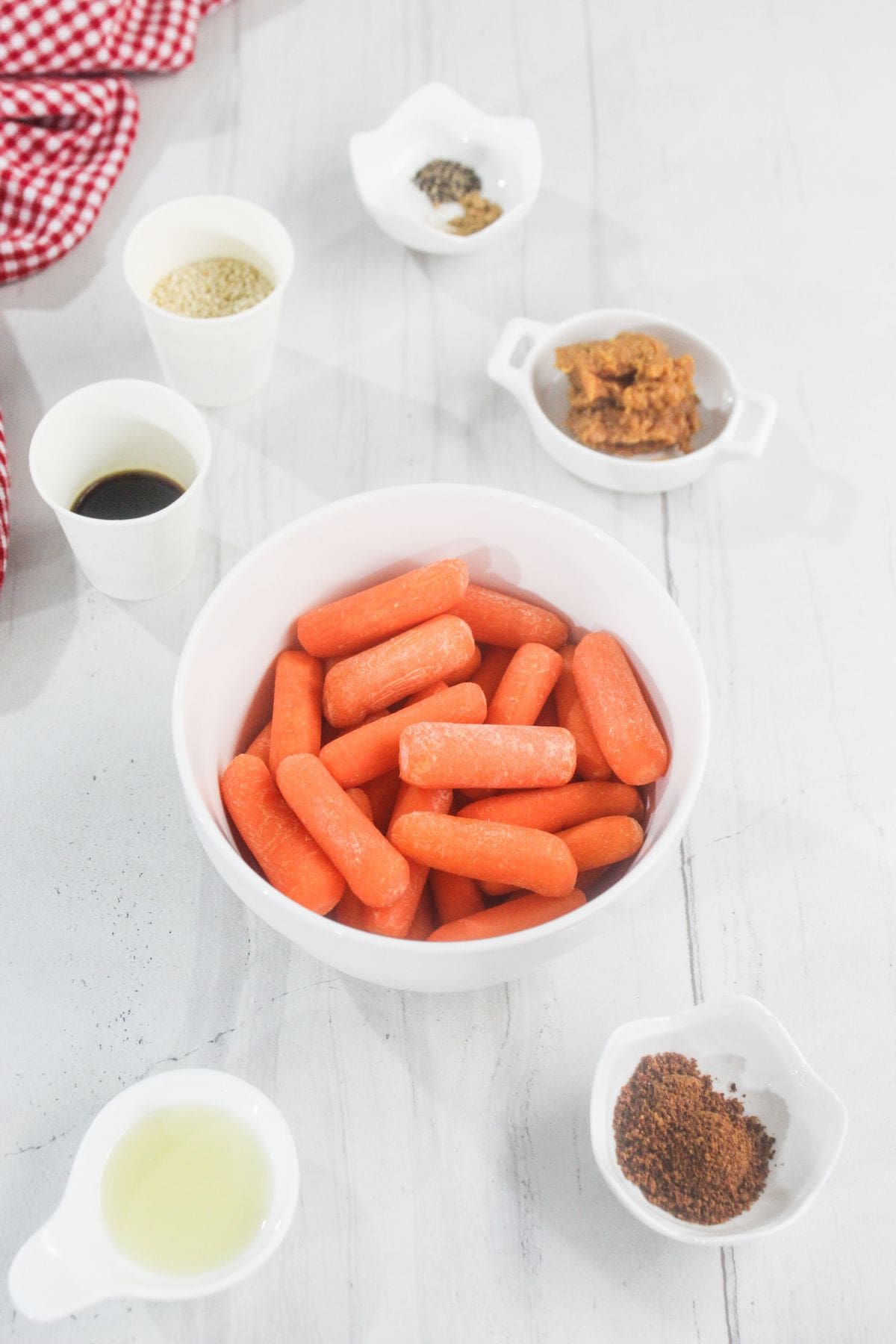 A bowl of baby carrots surrounded by small bowls containing seasonings, sauce, and condiments on a white surface.