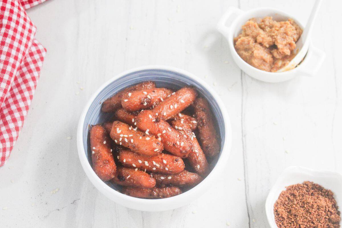 A bowl of glazed mini carrots topped with sesame seeds, next to bowls of brown seasoning and a chunky dipping sauce on a white surface.