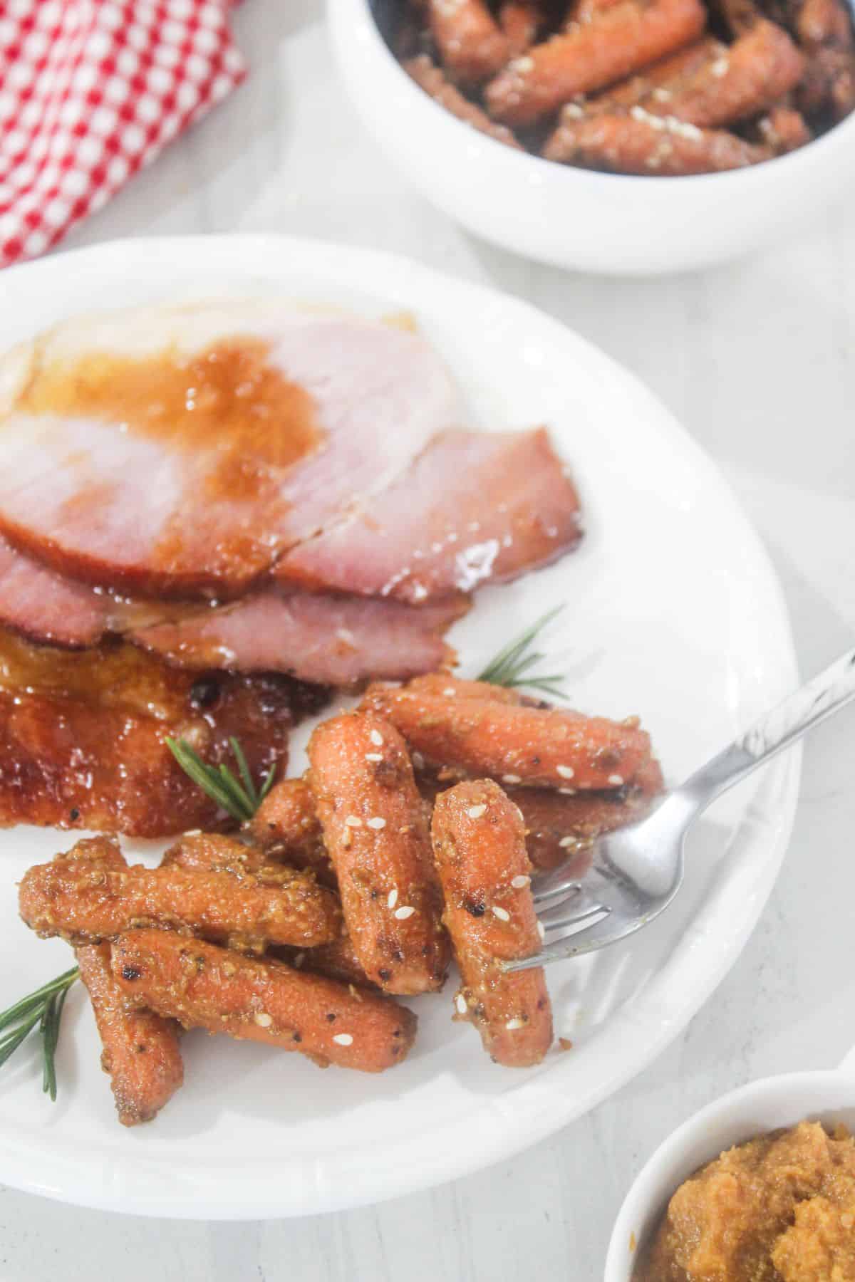 A white plate with sliced glazed ham and seasoned roasted baby carrots, garnished with rosemary. A fork holds some carrots. A small bowl of carrots and a red checkered napkin are nearby.