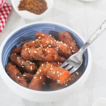 A bowl of glazed baby carrots topped with sesame seeds, with a fork resting inside the bowl.