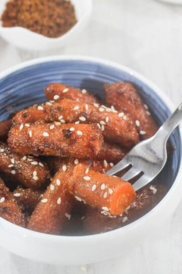 A bowl of glazed baby carrots topped with sesame seeds, with a fork resting inside the bowl.