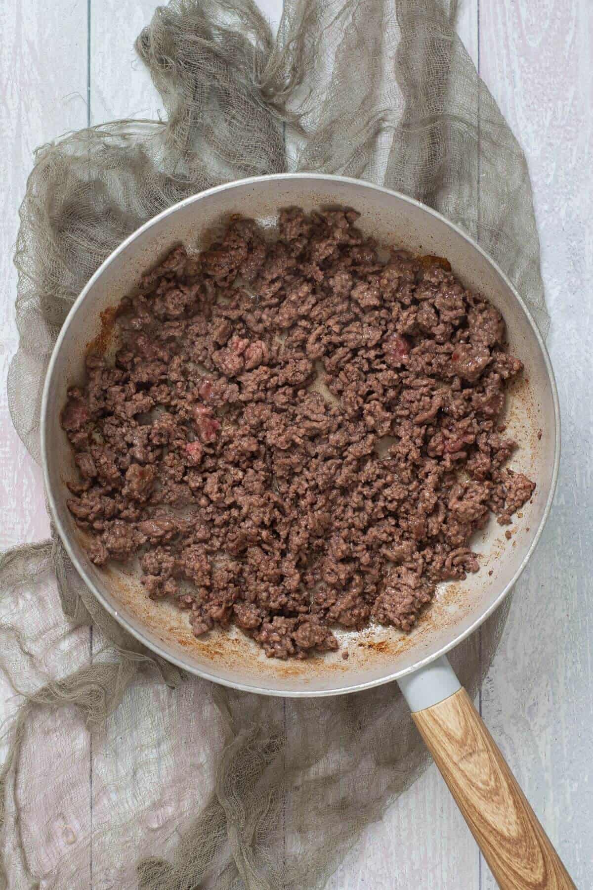 A frying pan with cooked ground beef on a light wooden surface, partially covered by a sheer, textured cloth.
