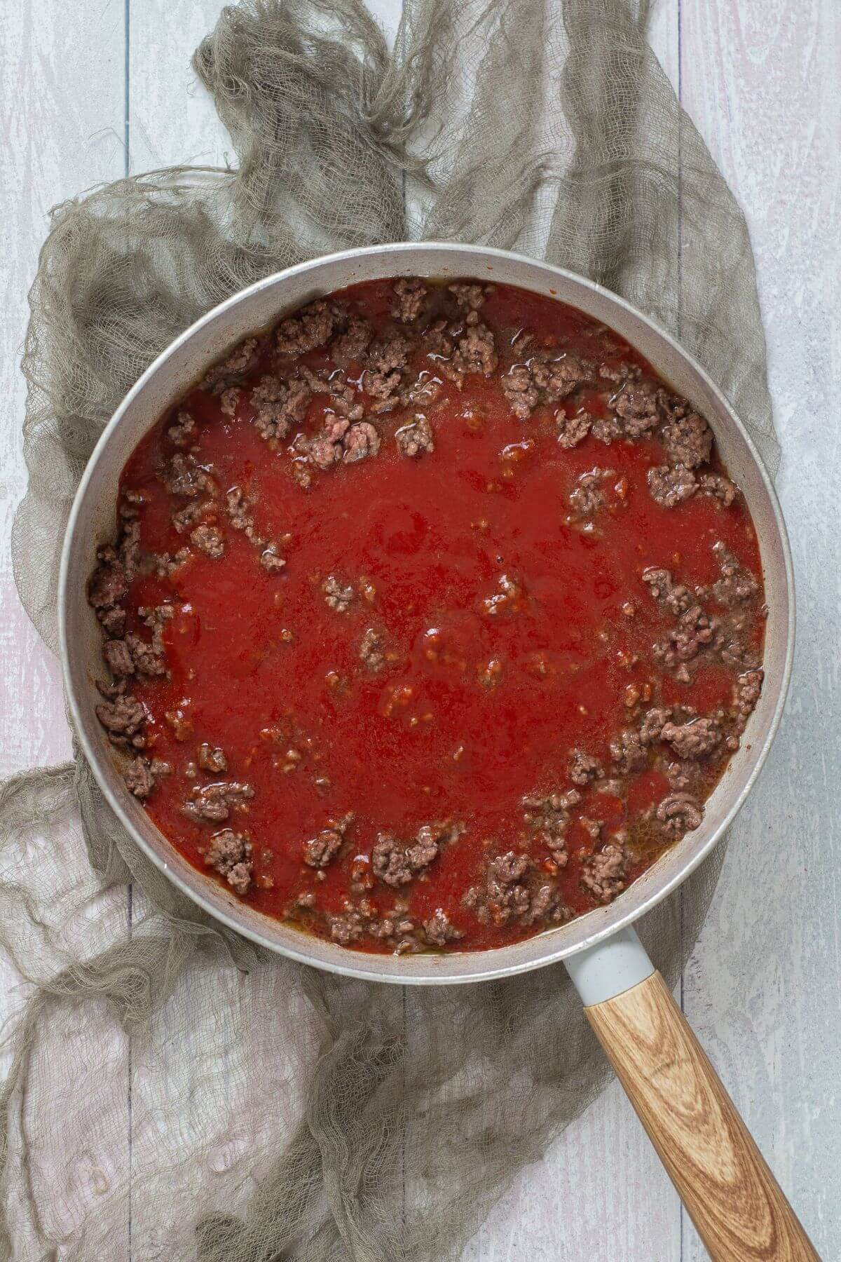 A pan with cooked ground beef and tomato sauce sits on a sheer cloth atop a white wooden surface.
