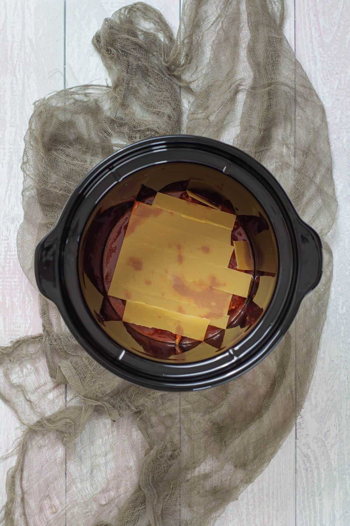 An overhead view of uncooked lasagna noodles layered over sauce in a black slow cooker, set on a light-colored wooden surface with a piece of beige fabric underneath.