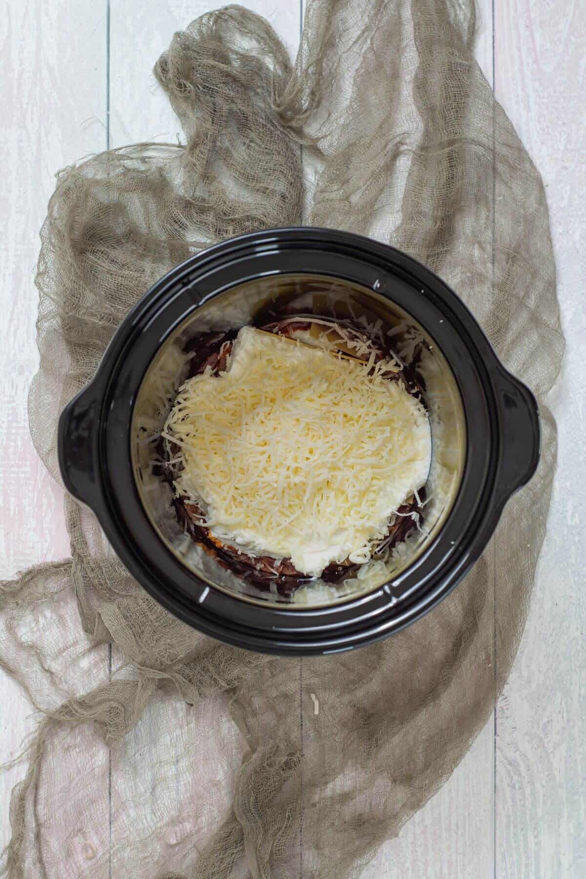 A slow cooker filled with layers of sauce, cheese, and possibly pasta, sits on a light wooden surface with a gray cloth underneath.