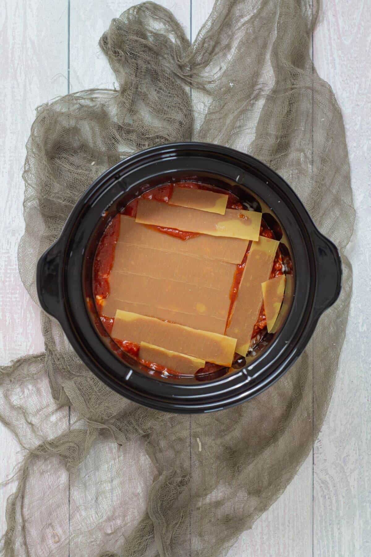 Top view of an uncooked lasagna in a slow cooker, with visible layers of dry lasagna noodles and tomato sauce.
