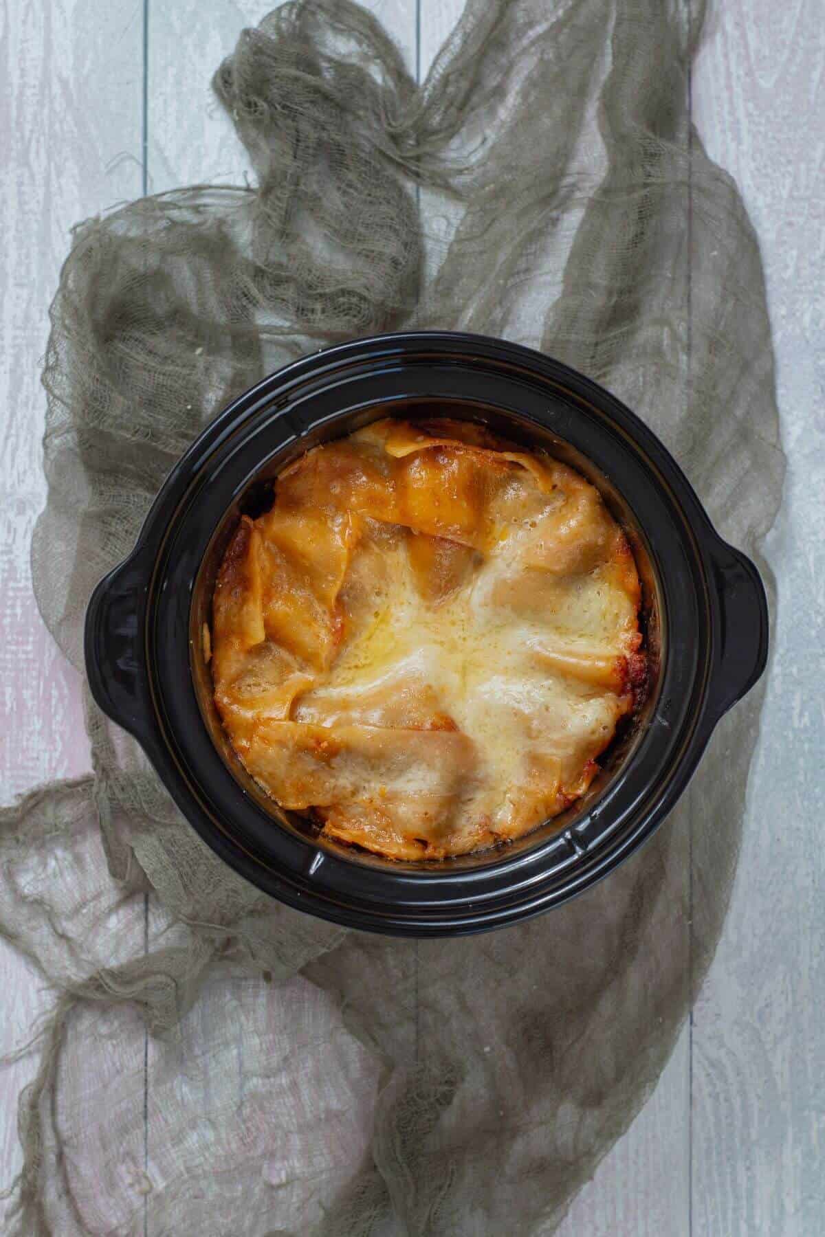 A round slow cooker filled with baked lasagna, topped with melted cheese, sits on a rustic wooden surface with a sheer gray cloth underneath.