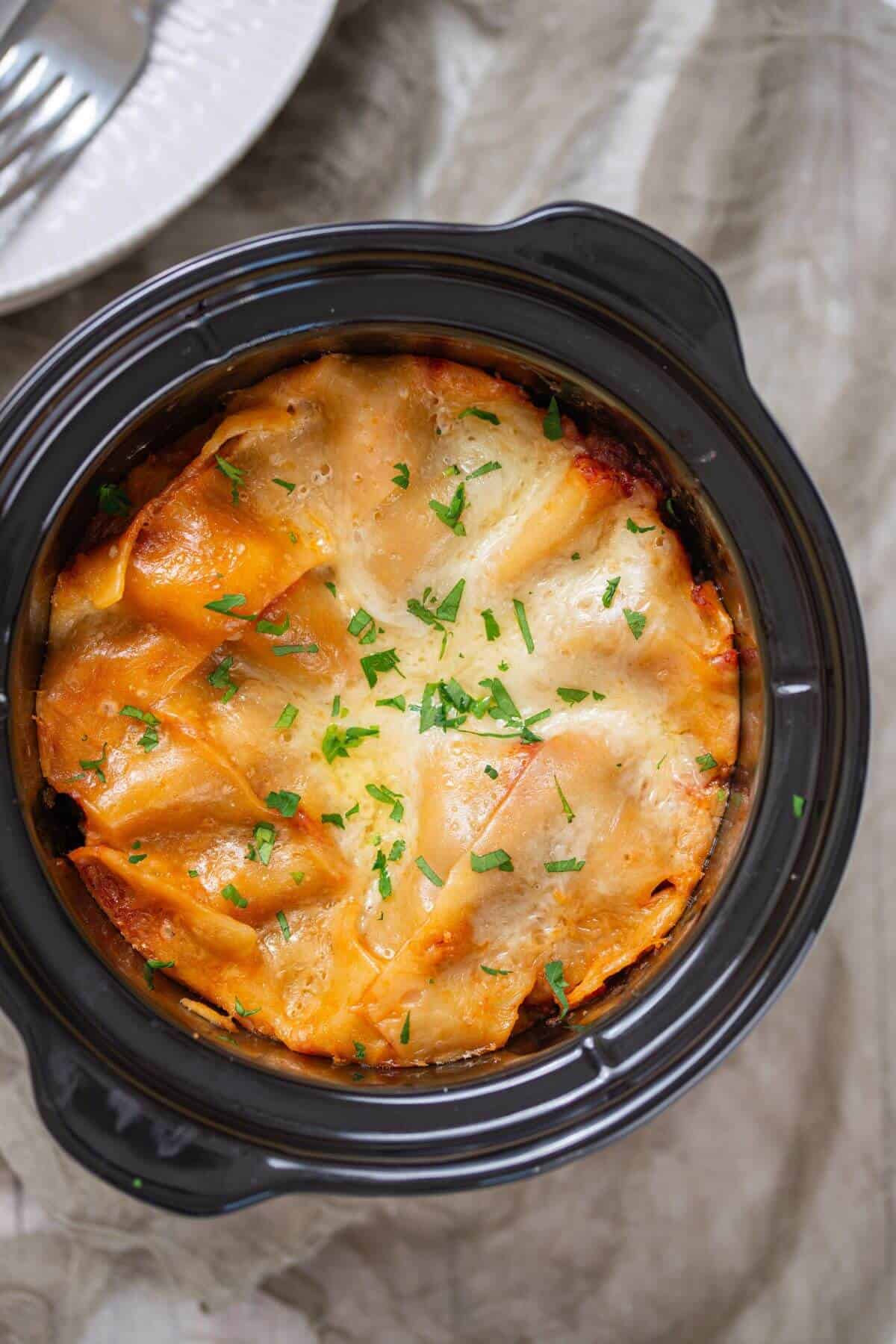 A crockpot filled with baked lasagna topped with melted cheese and sprinkled with chopped herbs on a beige cloth.