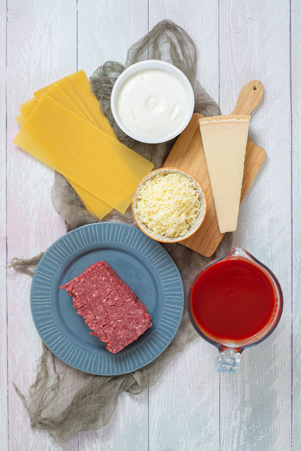 Top-down view of lasagna ingredients: uncooked lasagna noodles, ricotta cheese, parmesan, shredded mozzarella, ground beef, and tomato sauce arranged on a light wood surface.