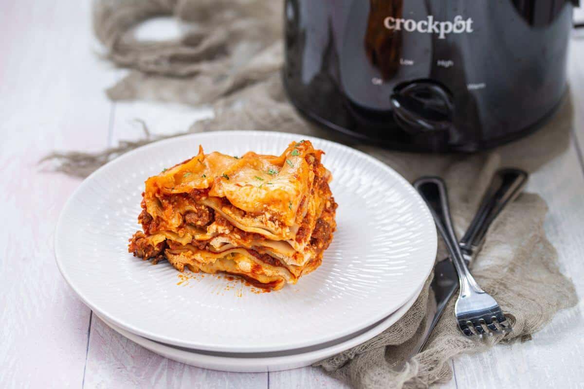 A slice of lasagna is served on a white plate next to a black Crock-Pot slow cooker and utensils on a napkin.