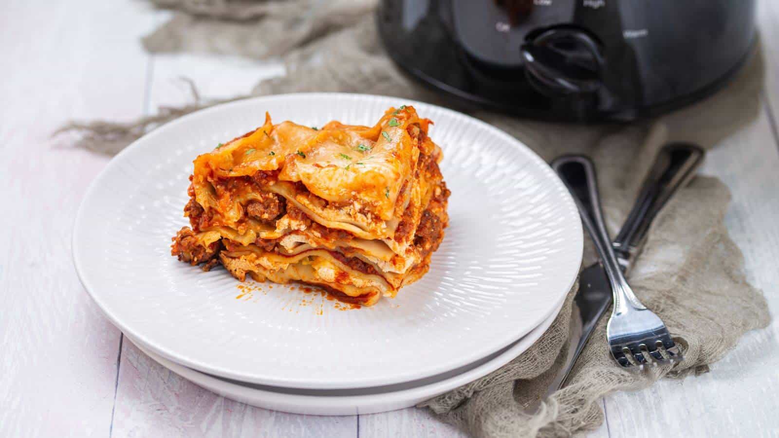 A serving of lasagna with layers of pasta, meat, and cheese sits on a white plate next to a black slow cooker and two utensils.