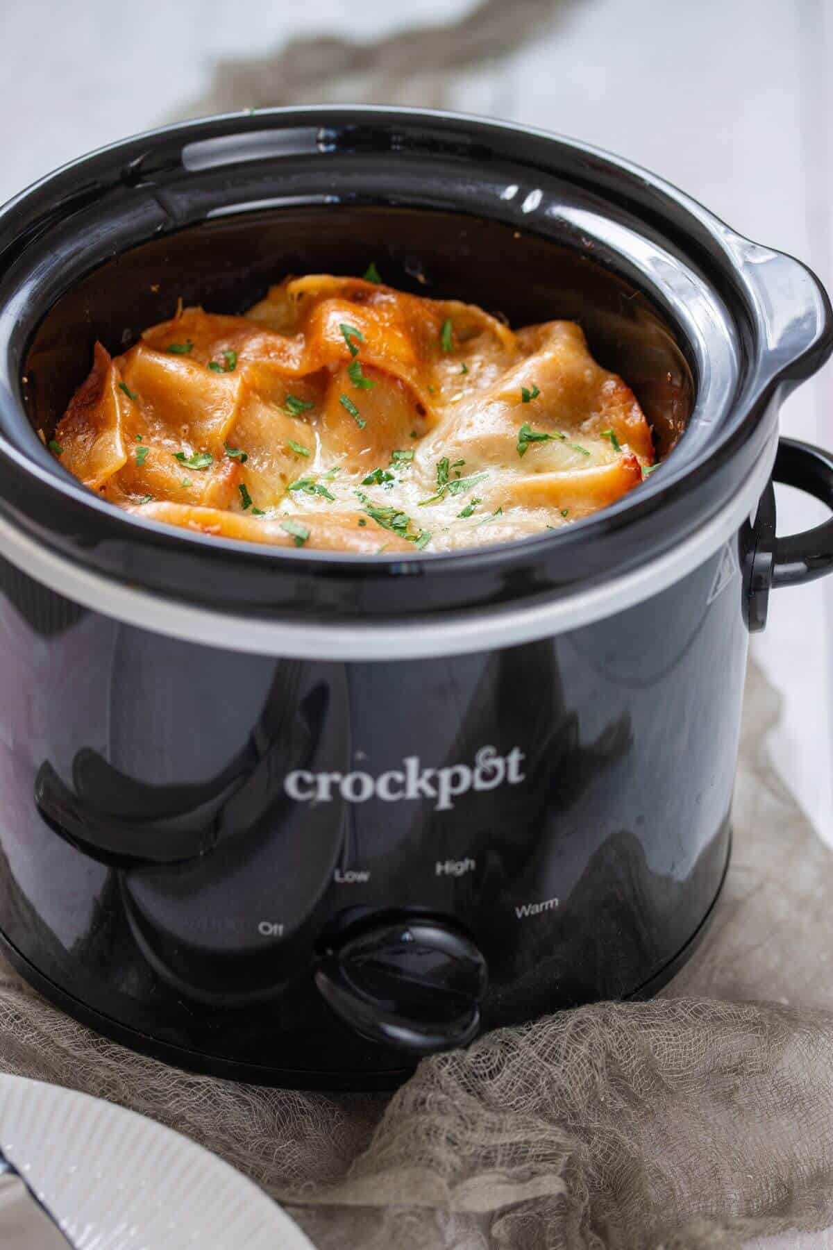 A black crockpot filled with baked lasagna, topped with melted cheese and chopped herbs, sits on a light-colored surface.
