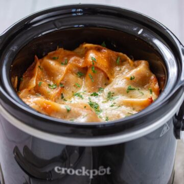 Lasagna with melted cheese and herbs is cooked in a black Crockpot slow cooker.