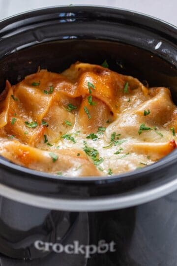 Lasagna with melted cheese and herbs is cooked in a black Crockpot slow cooker.