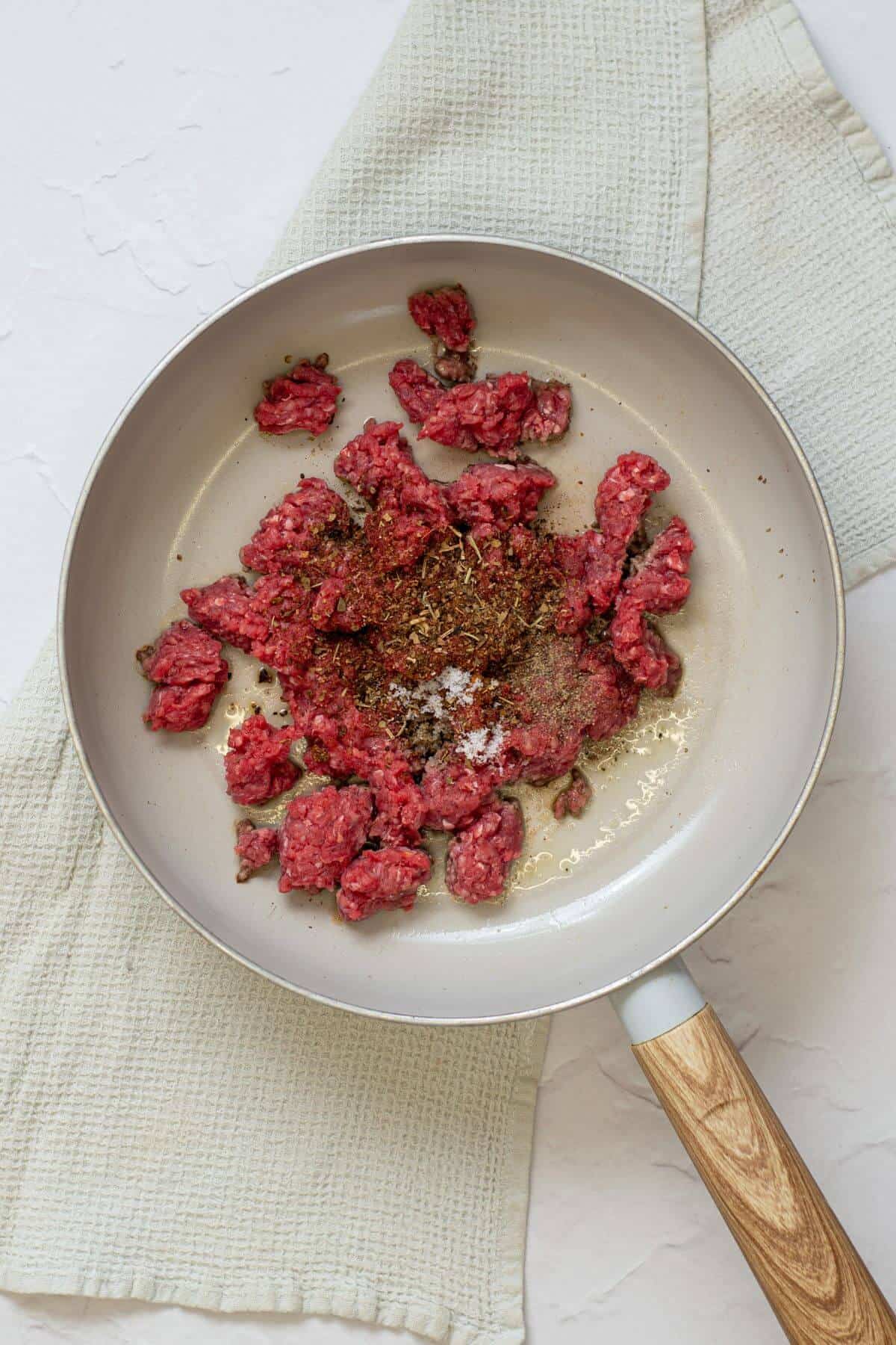 Ground beef and spices in a frying pan on a light cloth, ready to be cooked.