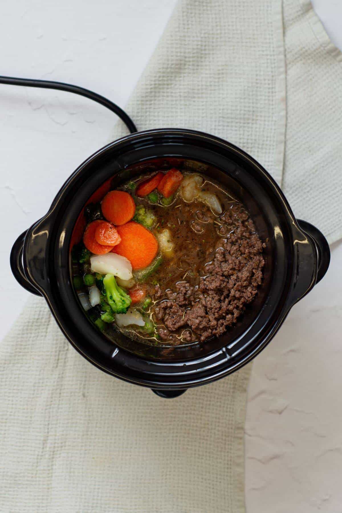 A slow cooker filled with ground beef, sliced carrots, chopped broccoli, onions, and broth, set on a light textured cloth.