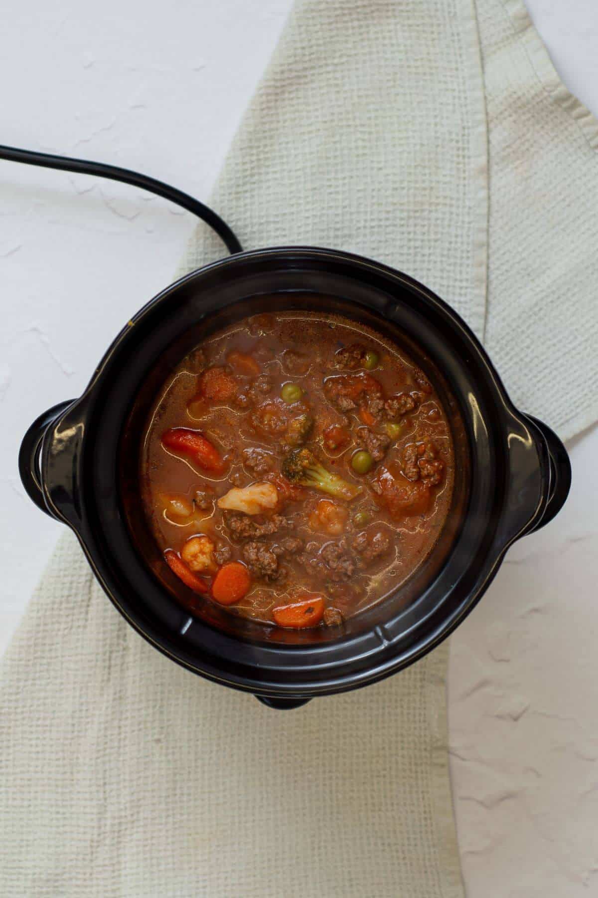 A black slow cooker filled with beef and vegetable stew sits on a light-colored textured cloth.
