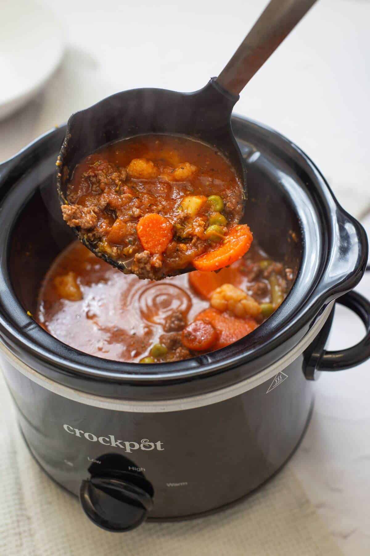 A black crockpot filled with beef stew containing carrots, peas, potatoes, and broth, with a ladle lifting a serving from the pot.