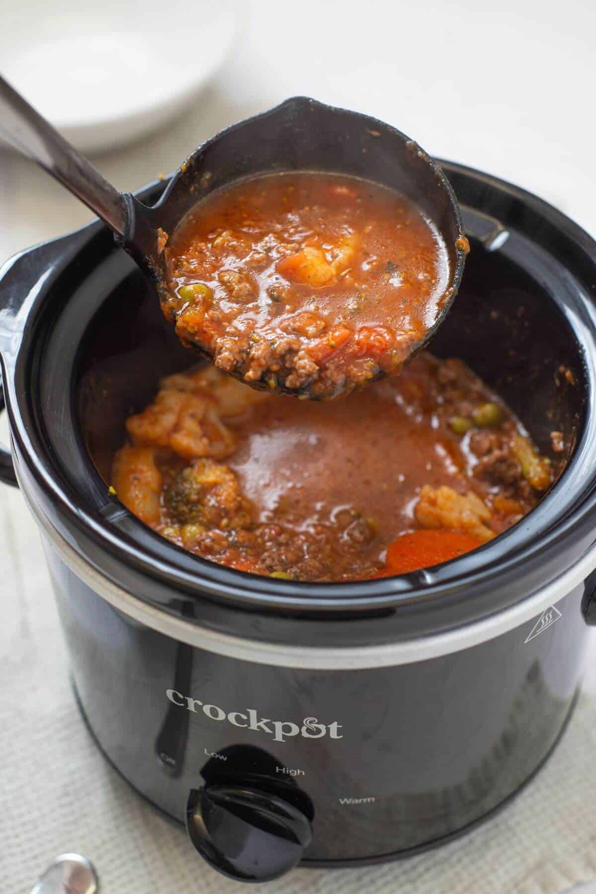 A ladle lifts a serving of chunky tomato-based stew from a black Crockpot slow cooker.
