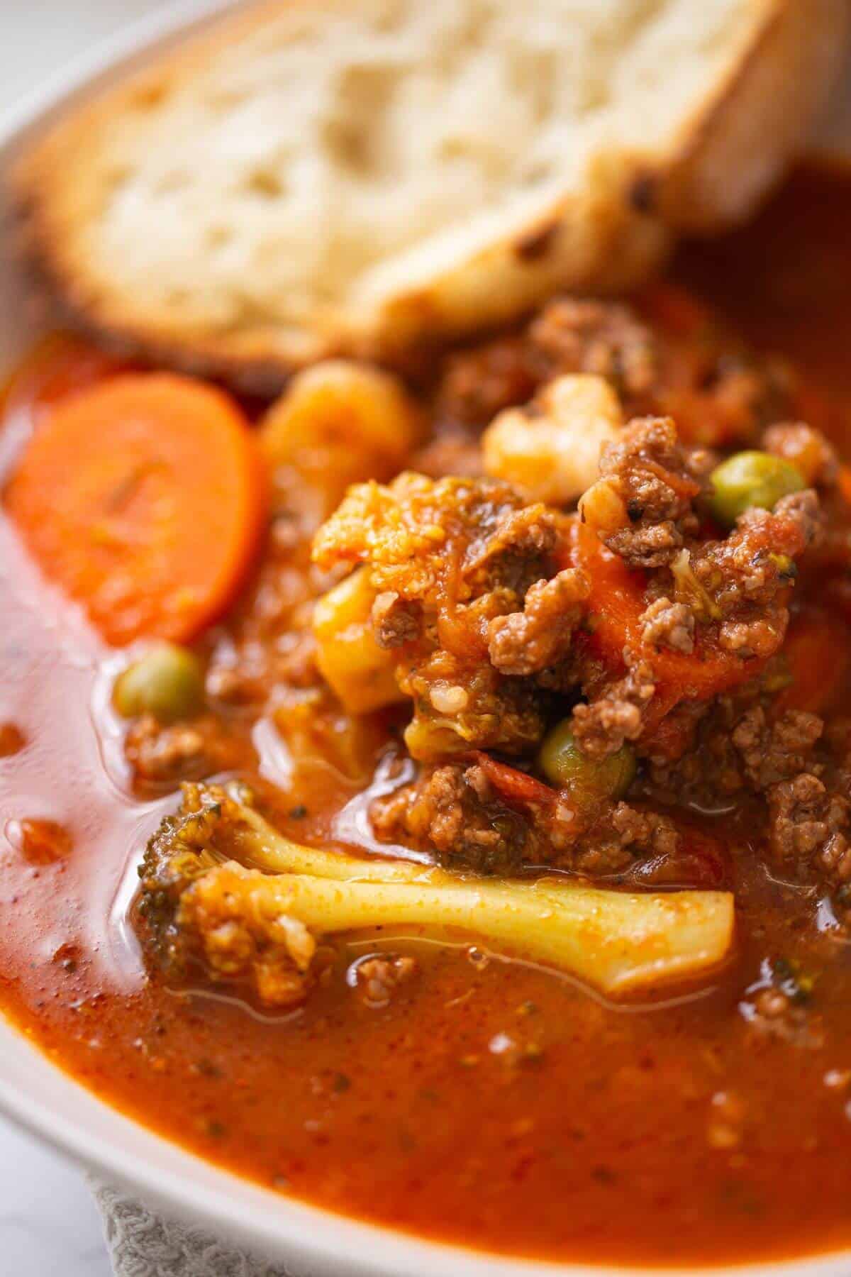 A bowl of chunky vegetable and ground beef soup with carrots, broccoli, peas, and a slice of bread on the side.