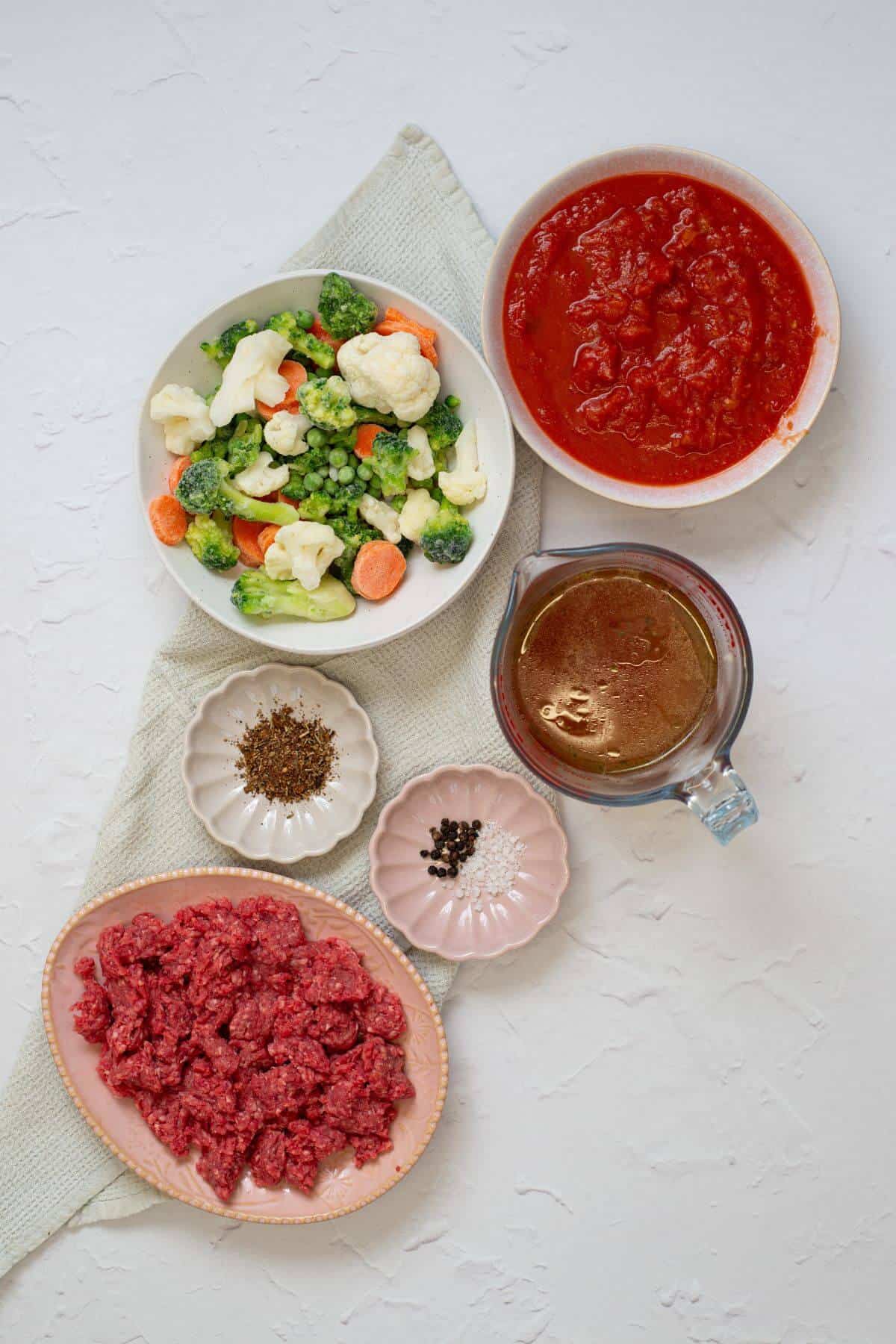 Bowls of frozen mixed vegetables, tomato sauce, ground beef, broth, and small plates of spices arranged on a white surface.