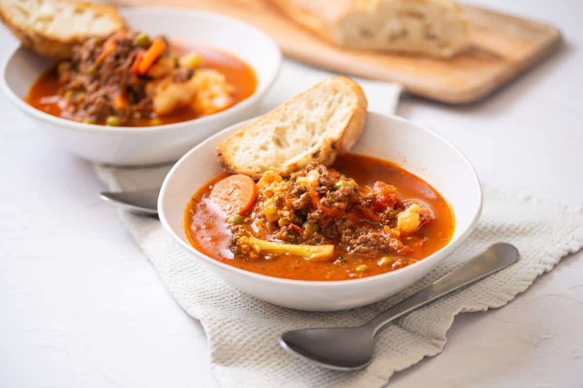 Two bowls of vegetable and beef soup with slices of bread, placed on a white cloth with spoons beside them; a loaf of bread is on a wooden board in the background.