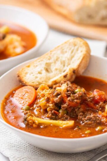 Two bowls of vegetable and ground meat soup are served with slices of bread on white plates, with a cutting board and more bread in the background.