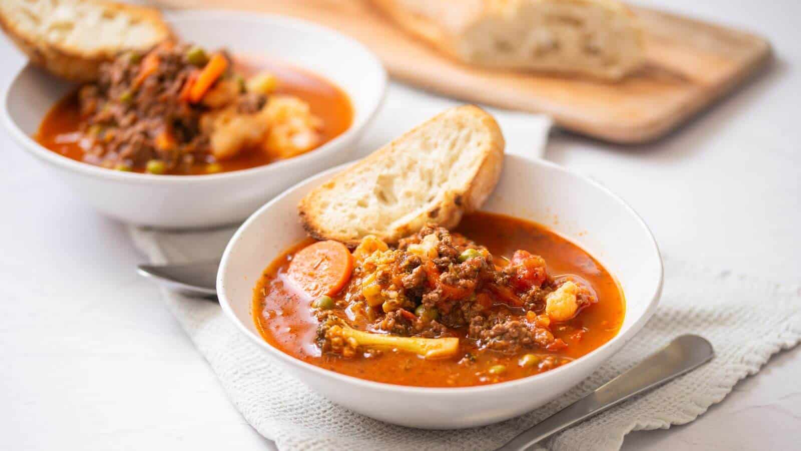 Two bowls of vegetable and ground meat soup are served with slices of bread on white plates, with a cutting board and more bread in the background.