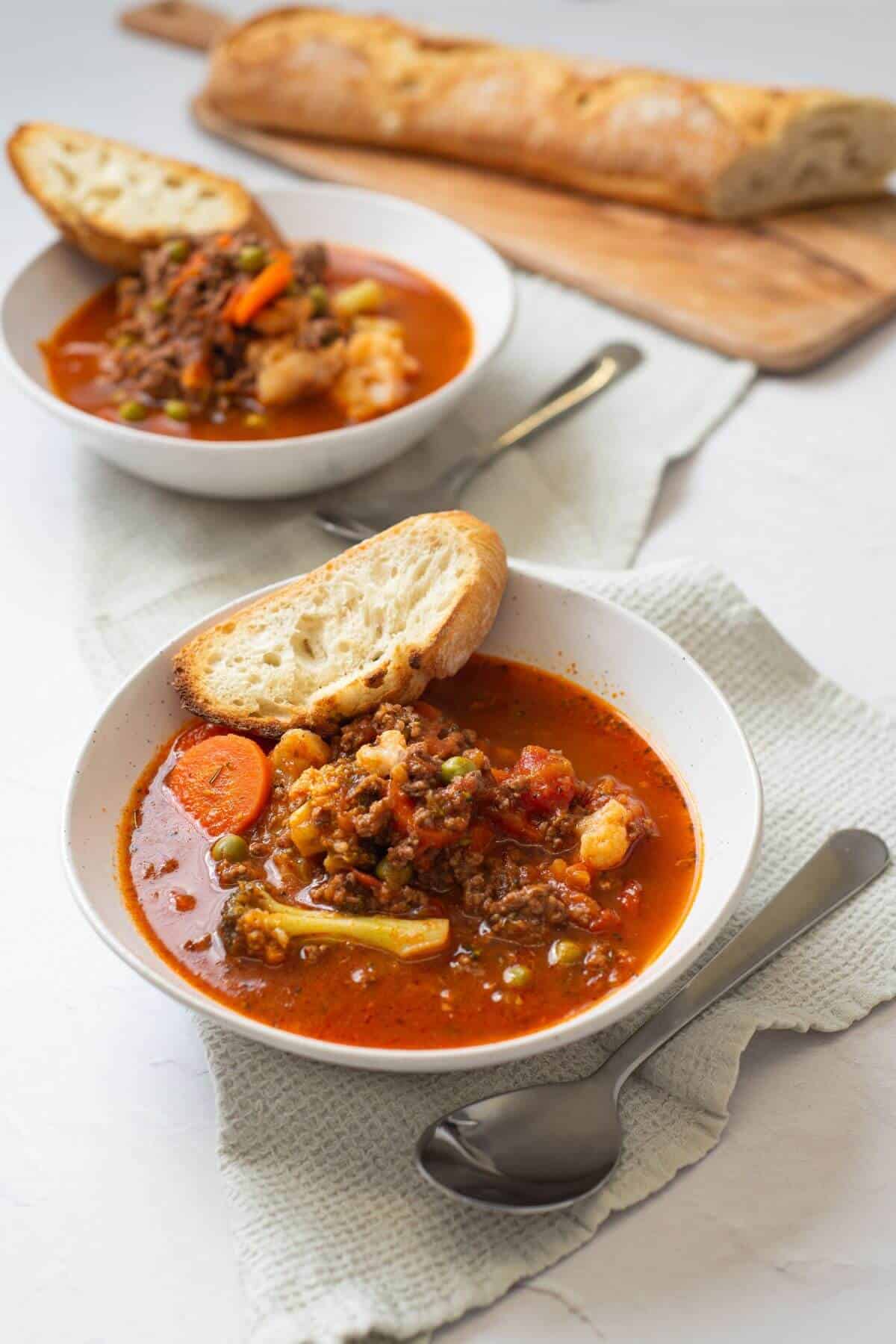 Two bowls of vegetable beef soup with bread slices, set on a light cloth with a baguette and knife in the background.