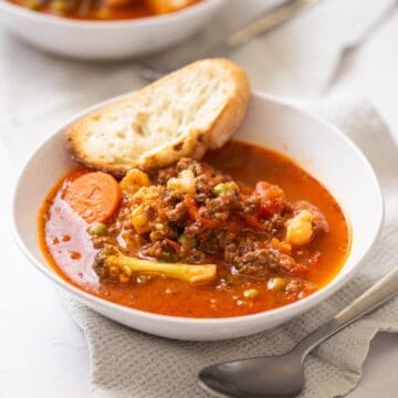 A bowl of vegetable and ground meat soup with a slice of bread resting on the edge, placed on a napkin with a spoon nearby.