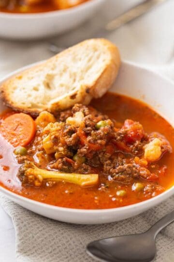 A bowl of vegetable and ground meat soup with a slice of bread resting on the edge, placed on a napkin with a spoon nearby.