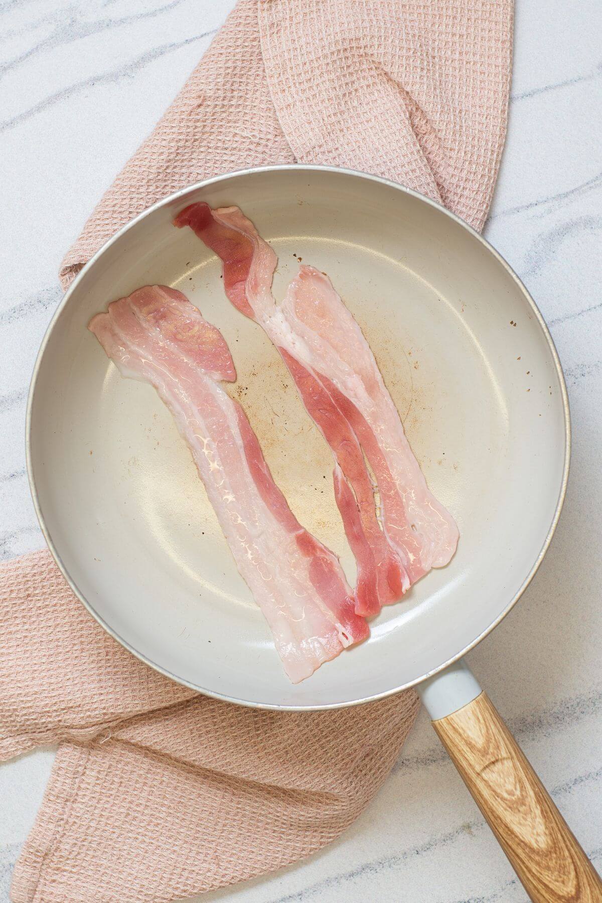 Two raw bacon strips in a light-colored frying pan placed on a pink textured towel on a white marble surface.