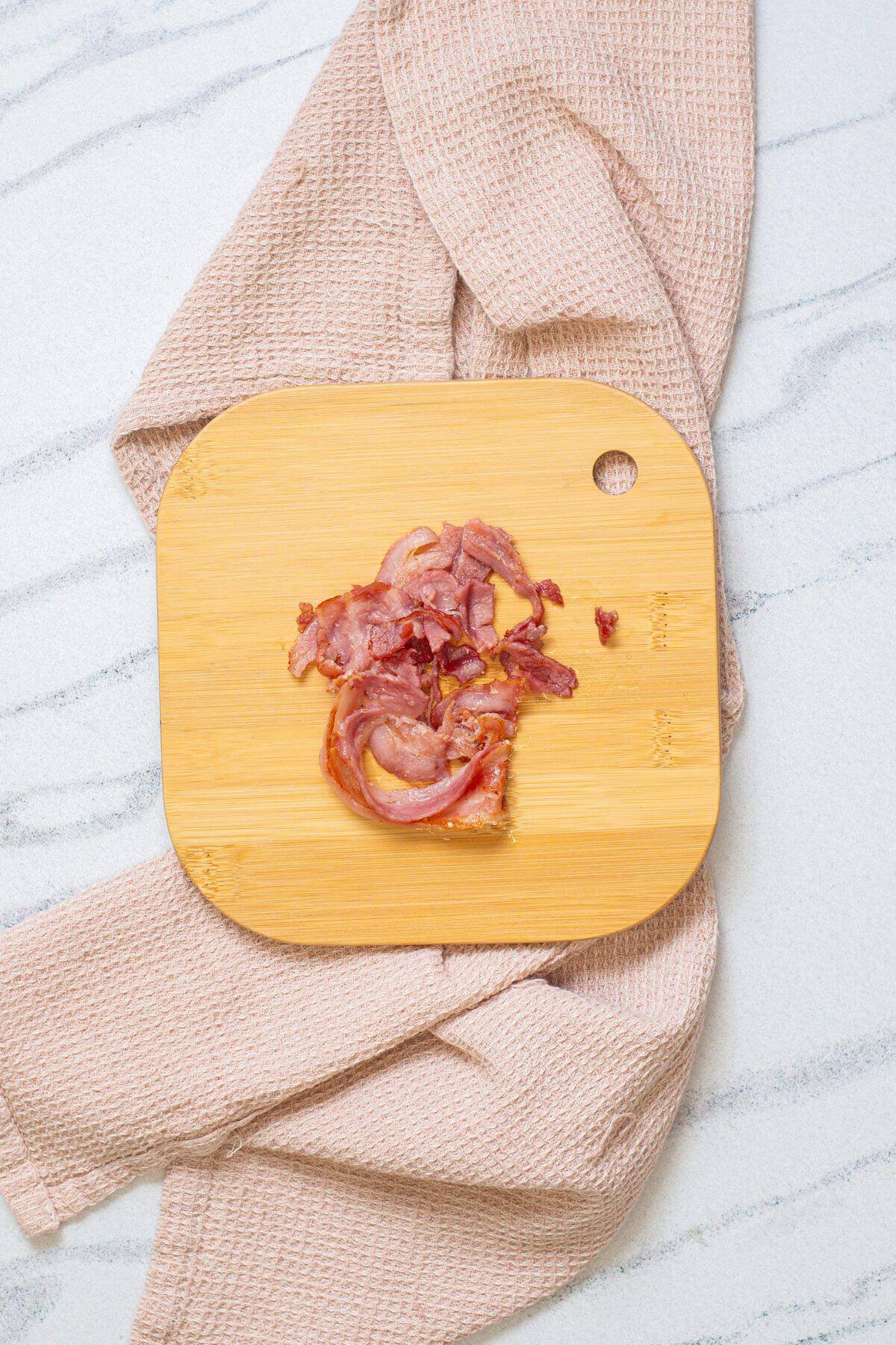 Chopped cooked bacon pieces arranged on a wooden cutting board placed on a textured beige cloth over a light-colored marble surface.