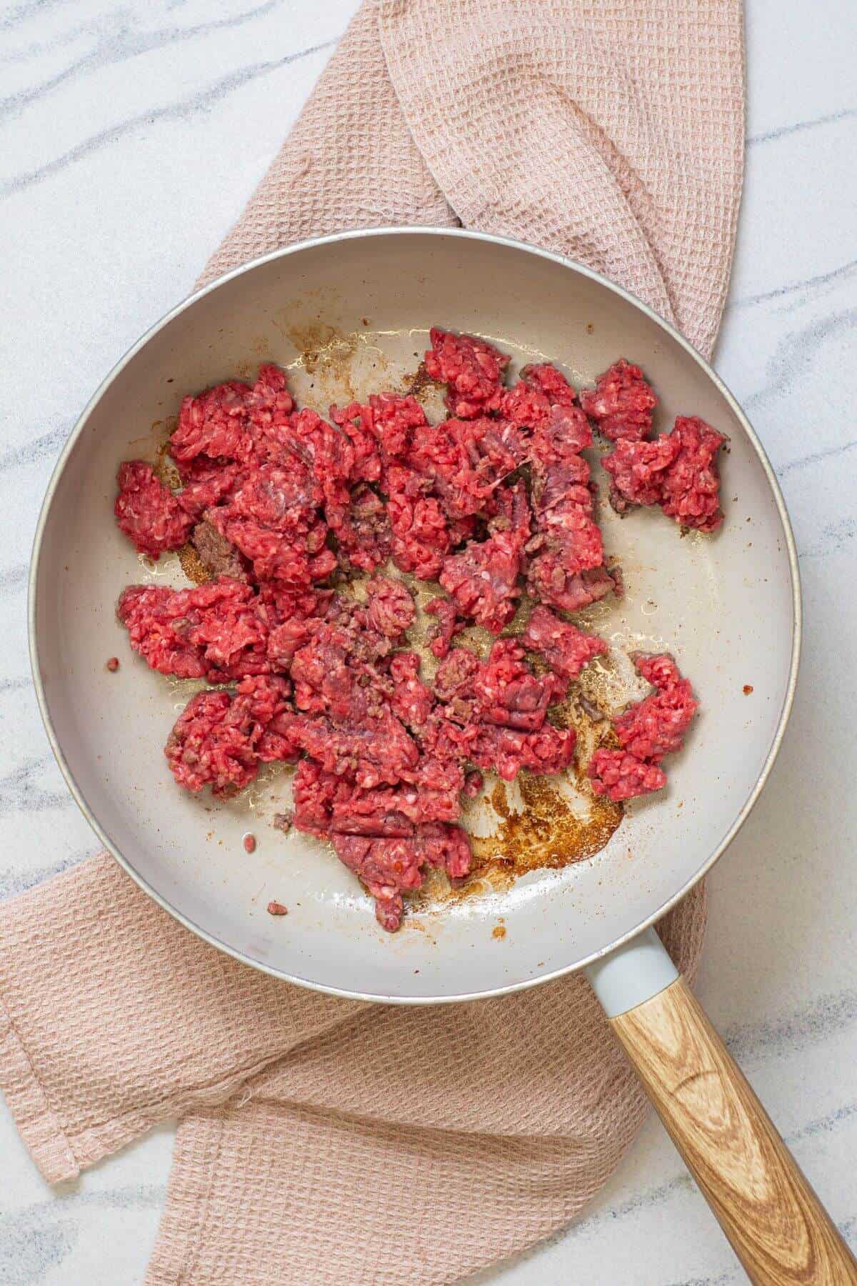 Ground beef cooking in a light-colored frying pan placed on a pink kitchen towel on a white countertop.