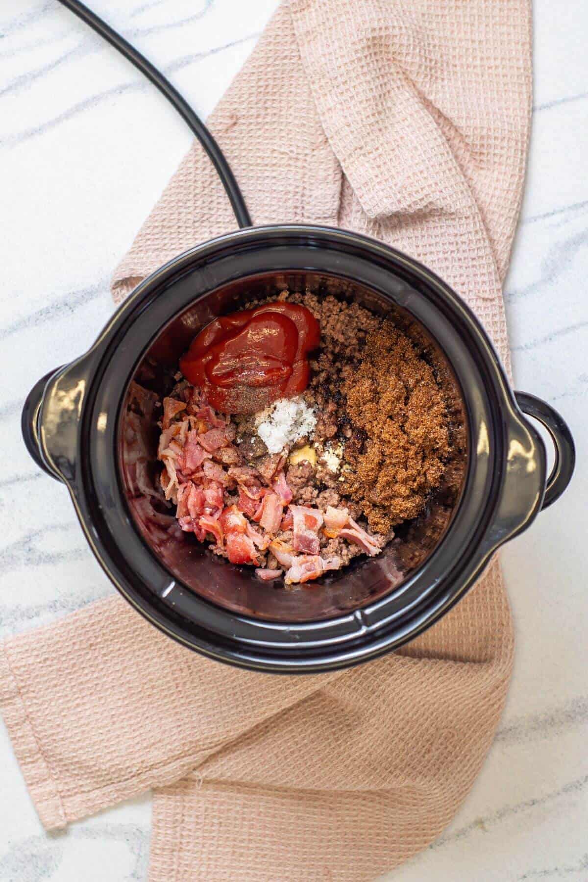 A crockpot filled with cooked ground beef, chopped bacon, tomato sauce, brown sugar, and seasonings, resting on a beige kitchen towel on a marble surface.